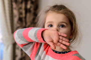 cute little brown-eyed girl in a striped pink-gray-white sweater wipes her nose with the back of...