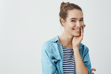 Girlfriend gazes at her couple with warm expression. Portrait of adorable european woman with bun hairstyle standing in trendy outfit over gray background, holding hand on cheek and smiling broadly