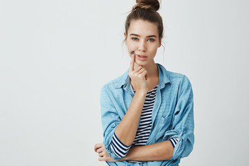 Fototapeta premium Woman is trying to think up where to put new sofa. Gorgeous modern woman with bun hairstyle holding hand on chin while looking with focused and determined expression at camera over gray background