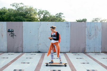 young woman standing on longboard and sending a text message from her cell phone. Woman using smart phone on skateboard outdoor © skvalval