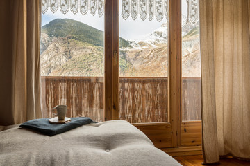 Vista de una habitación con un gran ventanal con vistas a unas impresionantes montañas cubiertas de nieve y sobre un sofá una taza blanca de café. 