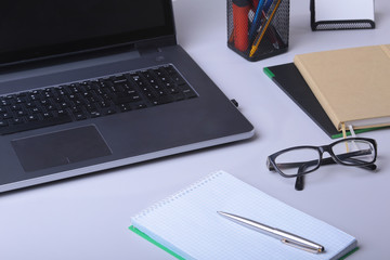 Close-up of comfortable working place in office with laptop, mouse, notebook, glasses, pen and other equipment laying on table on blur furniture background.