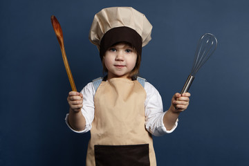 Naklejka premium Children, cooking, cuisine and food concept. Picture of funny little girl wearing chef uniform standing at blank wall, holding rolling pin in one hand and two wooden spoons in other, smiling at camera