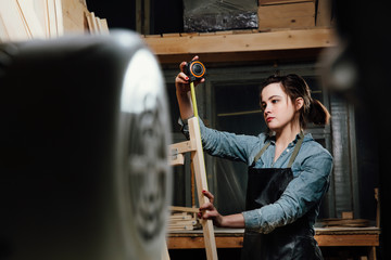 Fototapeta na wymiar Girl carpenter with a tape measure on a background of wooden pieces in workshop.