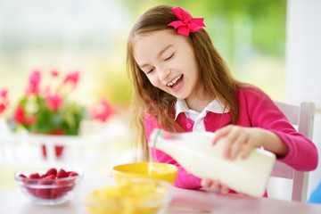 Cute little girl enjoying her breakfast at home. Pretty child eating corn flakes and raspberries and drinking milk before school. © MNStudio