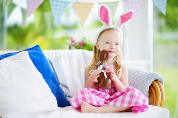 Cute little girl wearing bunny ears eating chocolate Easter rabbit. Kid playing egg hunt on Easter.