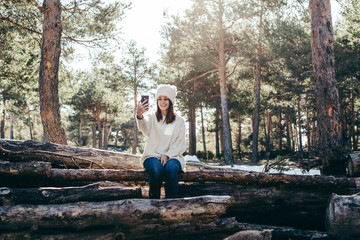 Fototapeta premium Young and pretty woman using her mobile phone outdoors, having fun taking herself pictures on a winter day with seated in a wooden trunk. Lifestyle.