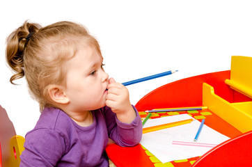 Little girl dreams and bites a pencil while sitting at a table, isolated on a white background