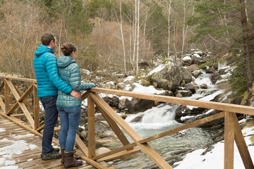 Pareja joven abrazados y mirando un río sobre un puente de madera. Paisaje invernal de montaña.