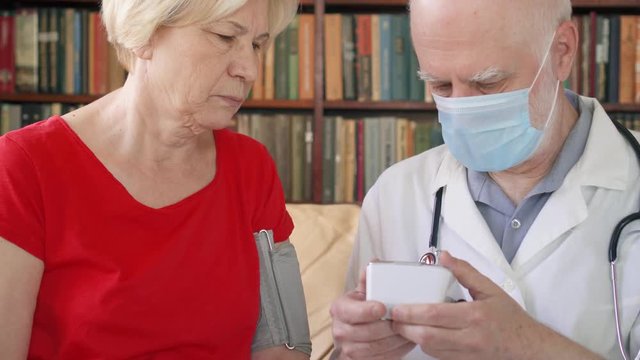 Male professional doctor in white coat and medical mask at work. Senior man physician measuring pressure to sick senior female patient by tonometer at home. Consulting about treatment and therapy