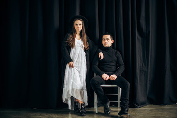 Stylish guy and girl on a black background, the guy is sitting on a chair and the girl is standing next to him in the hat on her head