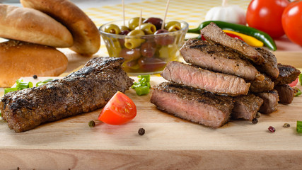 Grilled beef meat on wooden plate with vegetables and olives in the background.
