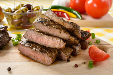 Grilled beef meat on wooden plate with vegetables and olives in the background.