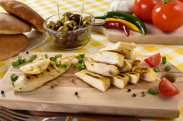Grilled chicken meat on wooden plate with vegetables and olives in the background.