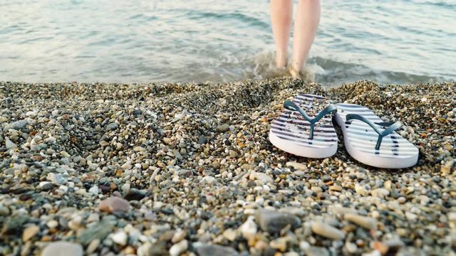 Woman feet red pedicure with blue striped flip flops summer shoes on sea shore stone beach. Holidays relax concept 4K