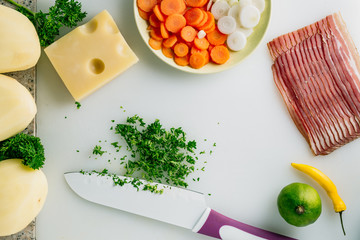Sliced parsley with knife, potato, cheese, bacon, some onion and herbs on table.