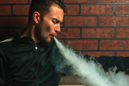 Vape man. Indoor portrait of a young handsome white guy letting of steam from an electronic cigarette. Vaping process. Close up.