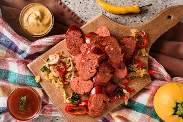 Sliced sausage and mix of fresh vegetables served on a rustic wooden board, mustard, ketchup, fresh hot chili peppers and herbs.
