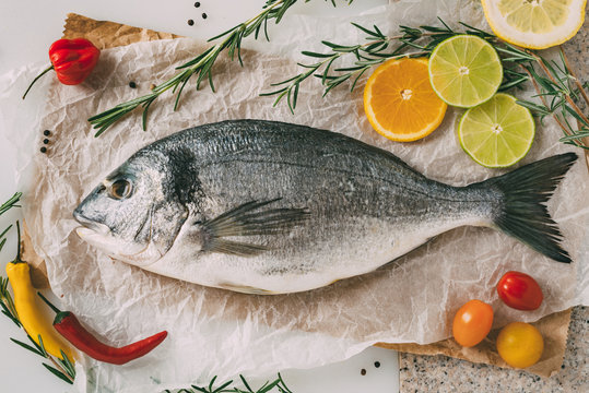 Sea gilt-head bream fish on baking sheet with rosemary, lemon, orange, tomato, hot peppers and lime. Fresh Orata, Dorade fish on kitchen table.