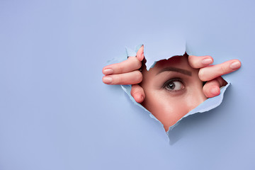 close-up of female fingers, nose and eye that can be seen through a hole in blue paper