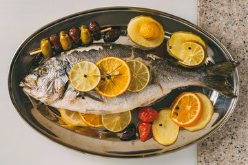 Sea gilt-head bream fish on the plate baked with potatoes, rosemary, lemon, orange, olives, tomatoes and lime. Fresh Orata, Dorade fish preparation.