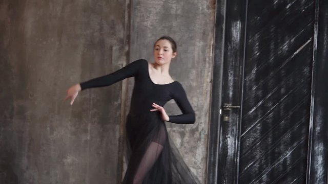 Pretty young woman practicing her dance show indoor alone. She is dressed in a black dress and looking elegant.