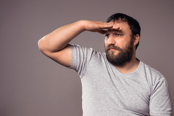 Bearded man in gray t-shirt looking away while keeping his hand over his eyes to protect from sun.