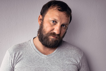 Handsome bearded man in gray t-shirt is looking at camera while standing against gray wall.