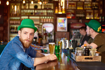 Portrait shot of handsome red-haired man wearing green bowler hat and denim shirt sitting at bar counter and enjoying fresh beer while celebrating St. Patricks Day with friends