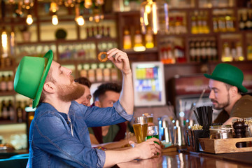 Profile view of bearded young man wearing green fedora hat and denim shirt sitting at bar counter, drinking fresh ale and eating appetizing snack, his friends chatting with each other