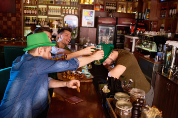 Group of cheerful friends toasting with beer glasses while sitting at bar counter and celebrating St. Patricks Day, bearded barman wearing green bowler hat putting his head under glasses
