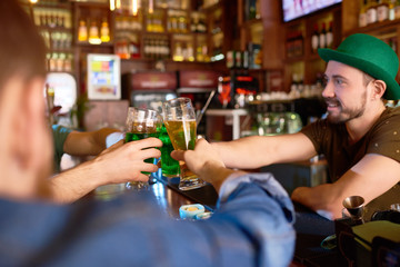 Group of cheerful friends toasting with beer glasses while celebrating St. Patricks Day in modern pub, handsome bearded barman wearing green bowler hat