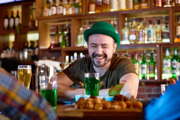 Head and shoulders portrait of cheerful bearded barman wearing green bowler hat leaning on bar counter and laughing while having fun with group of visitors