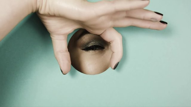 Girl's eye with beautiful make-up,with sparkles,with shadows, peeps through circular crevice of colored paper. The girl playfully peeping through hole. Her palm makes binoculars. Concept advertising.