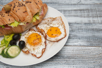 White Plate with Fried Eggs and Croissant Sandwich with Cucumber, Tomatoe and Olives. Breakfast. Gray Wooden Background. Rustic Style with copy Paste.