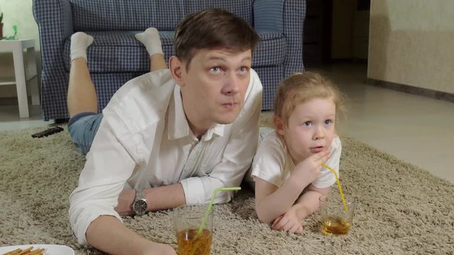 man and daughter watching television, sitting on the floor drink juice