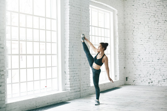 Full lenght image of slim beautiful young woman with muscular body exercising indoors, doing standing splits by large window. Flexible girl stretching legs in spacious gym hall at fitness center