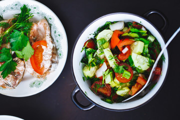 Fresh vegetable salad in plate and fish on black background, close up, top view. Healthy food, lunch.