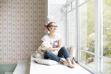Portrait of childish beautiful pink haired Caucasian female teenager wearing trendy black eyeglasses, choker and no socks hugging her knitted rabbit toy, relaxing on windowsill, enjoying leisure time