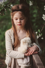 cute dreamy child girl posing at rustic wooden fence with teddy bear and blooming cherry on background