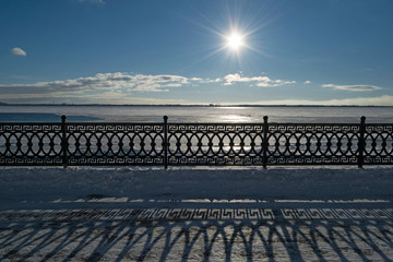 The sun over the winter river casts long shadows from the beautiful fence. Decorative fencing and embankment of the Volga River in winter. Quay zone in the city of Saratov.