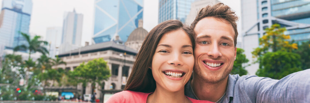 Young smiling people selfie portrait panorama banner. Two tourist couple traveling in Hong Kong city taking photo with phone in summer vacation. Asia travel lifestyle. Asian girl, Caucasian man.