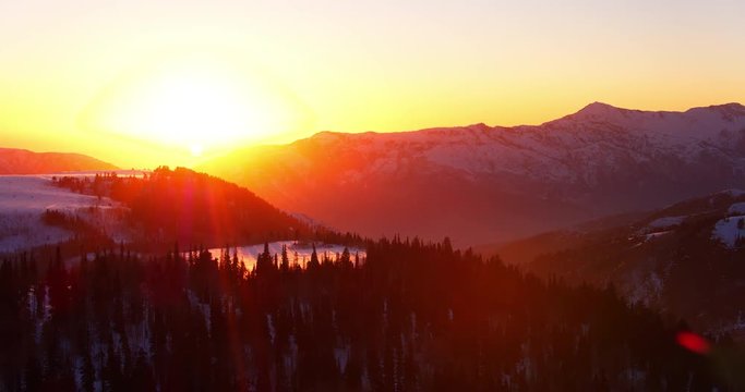 Aerial view flying over winter forest at sunset with snow covered mountains in background