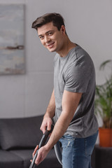 handsome man cleaning carpet with vacuum cleaner and looking at camera in living room