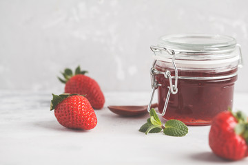 Strawberry jam in a glass jar with berries, gray background.