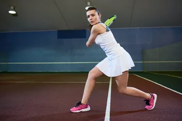 Tuinposter Full length portrait of confident young woman playing tennis in indoor court, ready to hit flying ball, copy space © Seventyfour