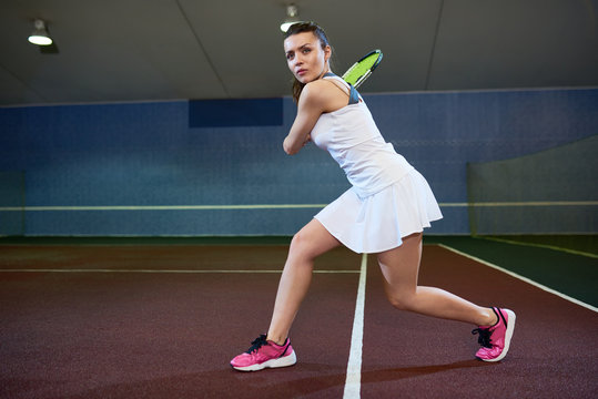 Full length portrait of confident young woman playing tennis in indoor court, ready to hit flying ball, copy space