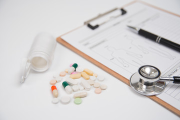 Pills and a white bottle, along with a stethoscope and a medical chart, are over a white background. Used for health or medical manufacturing industry concept.