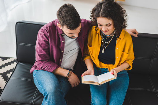 High Angle View Of Young Couple Sitting On Couch And Reading Book