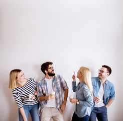 Group of young handsome colleague people standing against the wall having fun and drinking coffee in paper cups for a break while one girl showing up with a finger on empty editable space.